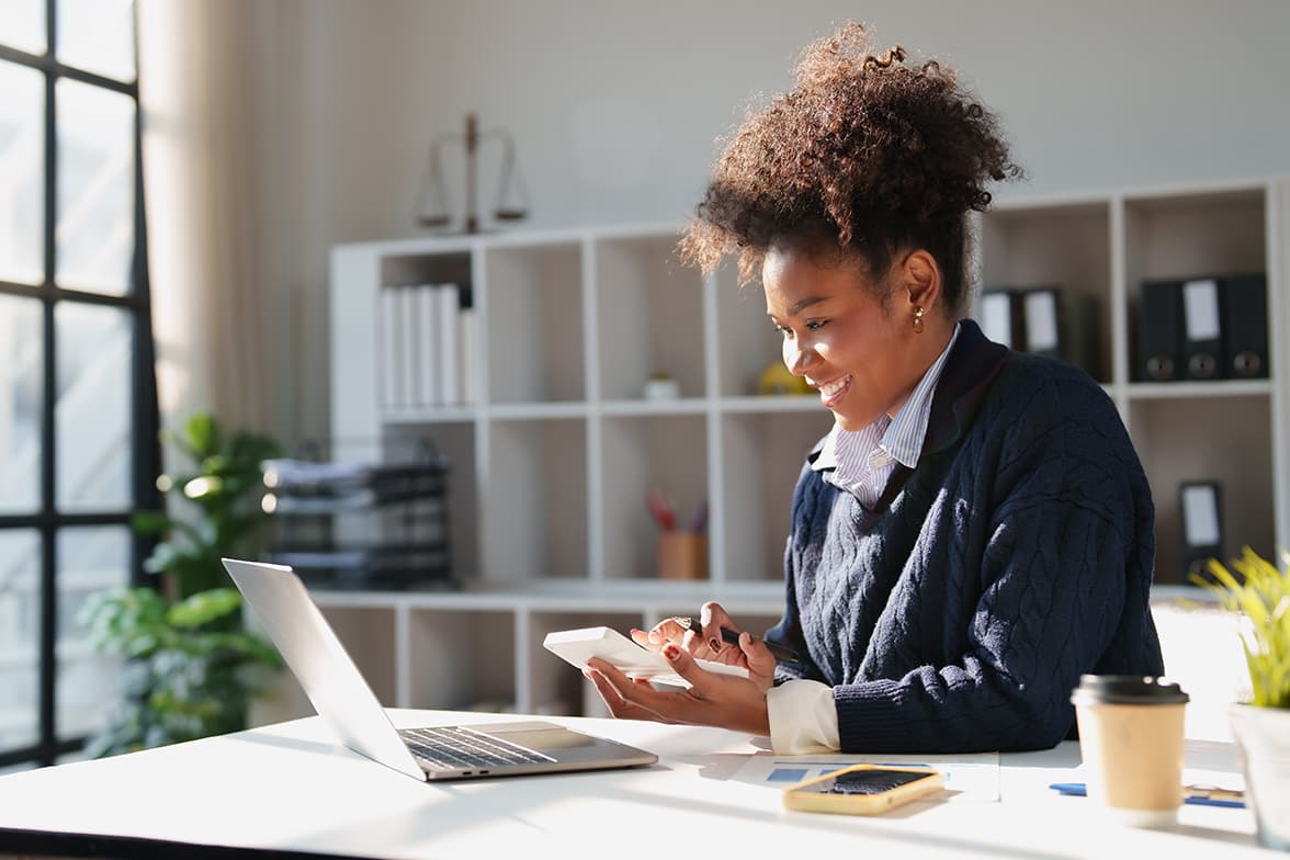 A woman siting at a desk using a calculator while looking at a laptop.