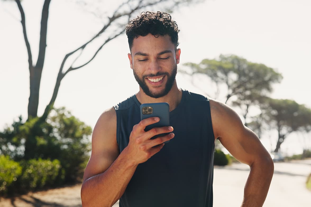 Smiling man in athletic wear looking at his smartphone outdoors on a sunny day.